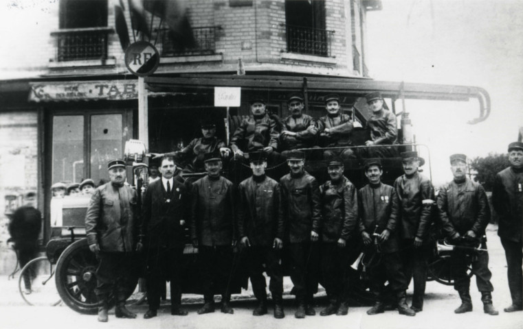 Pompiers posant devant leur véhicule motorisé garé devant un café : reproduction de photographie noir et blanc.