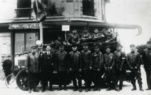 Pompiers posant devant leur véhicule motorisé garé devant un café : reproduction de photographie noir et blanc.