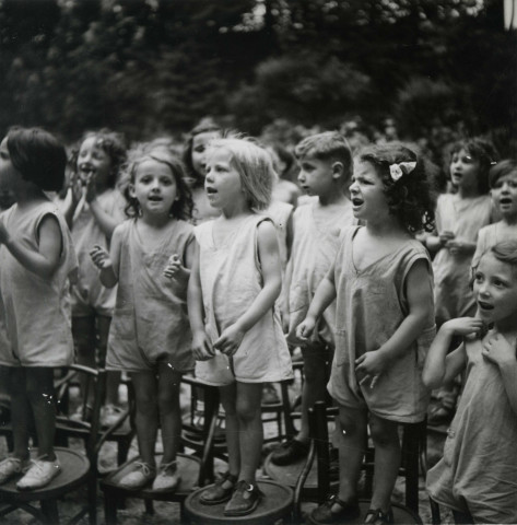 École de Plein-Air, groupe d'enfants chantant debout sur des chaises en extérieur : photographie noir et blanc.