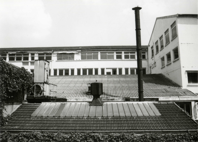Usine Motobécane désaffectée, vue de l'extérieur sur les toitures et une partie des bâtiments : photographie noir et blanc.