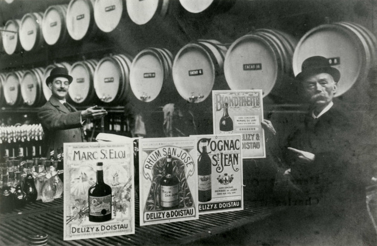 Affiches publicitaires de la distillerie Delizy et Doistau présentées par deux hommes devant des tonneaux : reproduction d'une photographie noir et blanc.