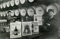 Affiches publicitaires de la distillerie Delizy et Doistau présentées par deux hommes devant des tonneaux : reproduction d'une photographie noir et blanc.